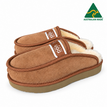 Load image into Gallery viewer, A pair of brown suede slip-on slippers with white shearling lining and a white UGG logo tab, shown on a clean white background.